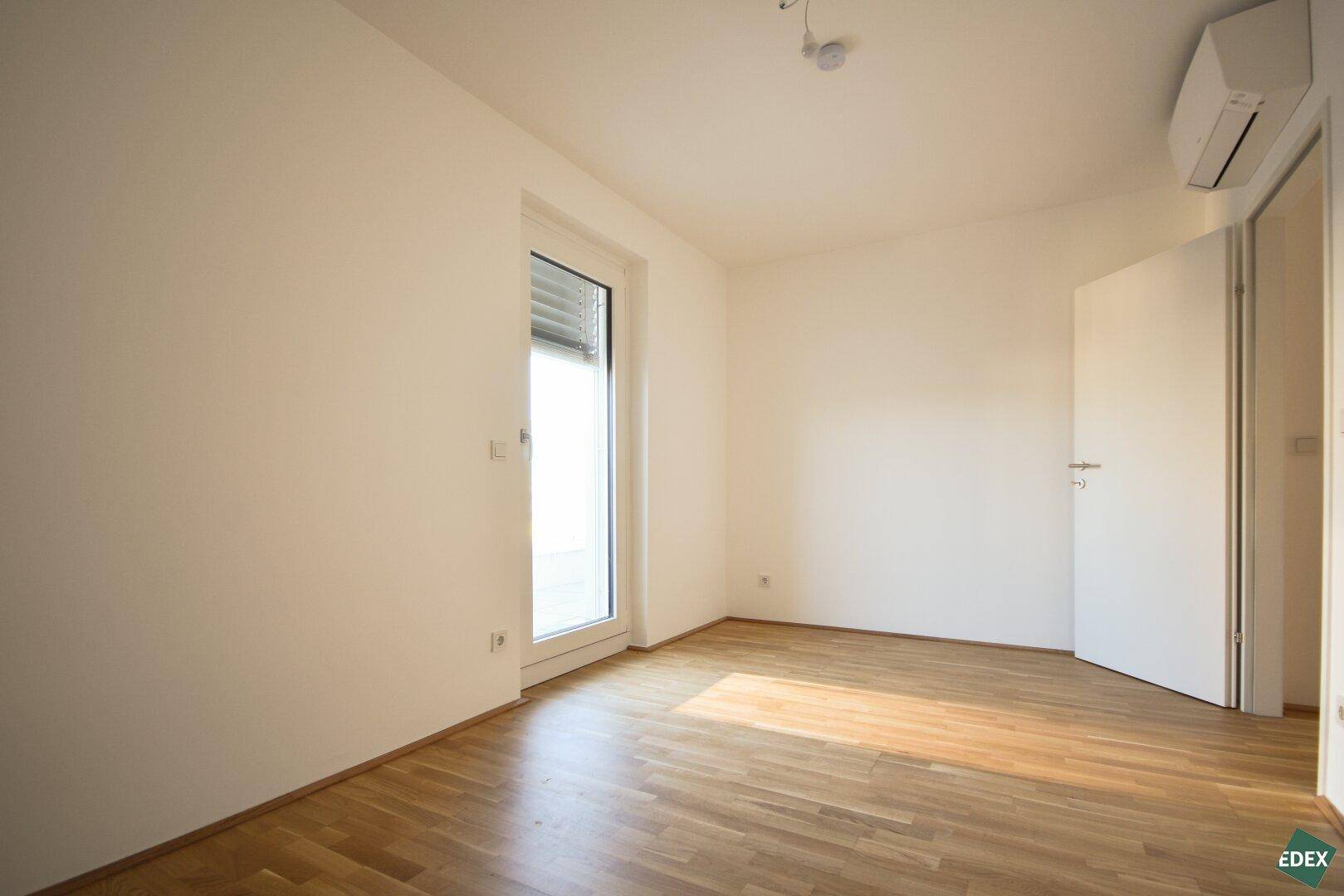 Leeres Schlafzimmer mit Holzboden, Klimaanlage und Balkonzugang, bereit zur Einrichtung.