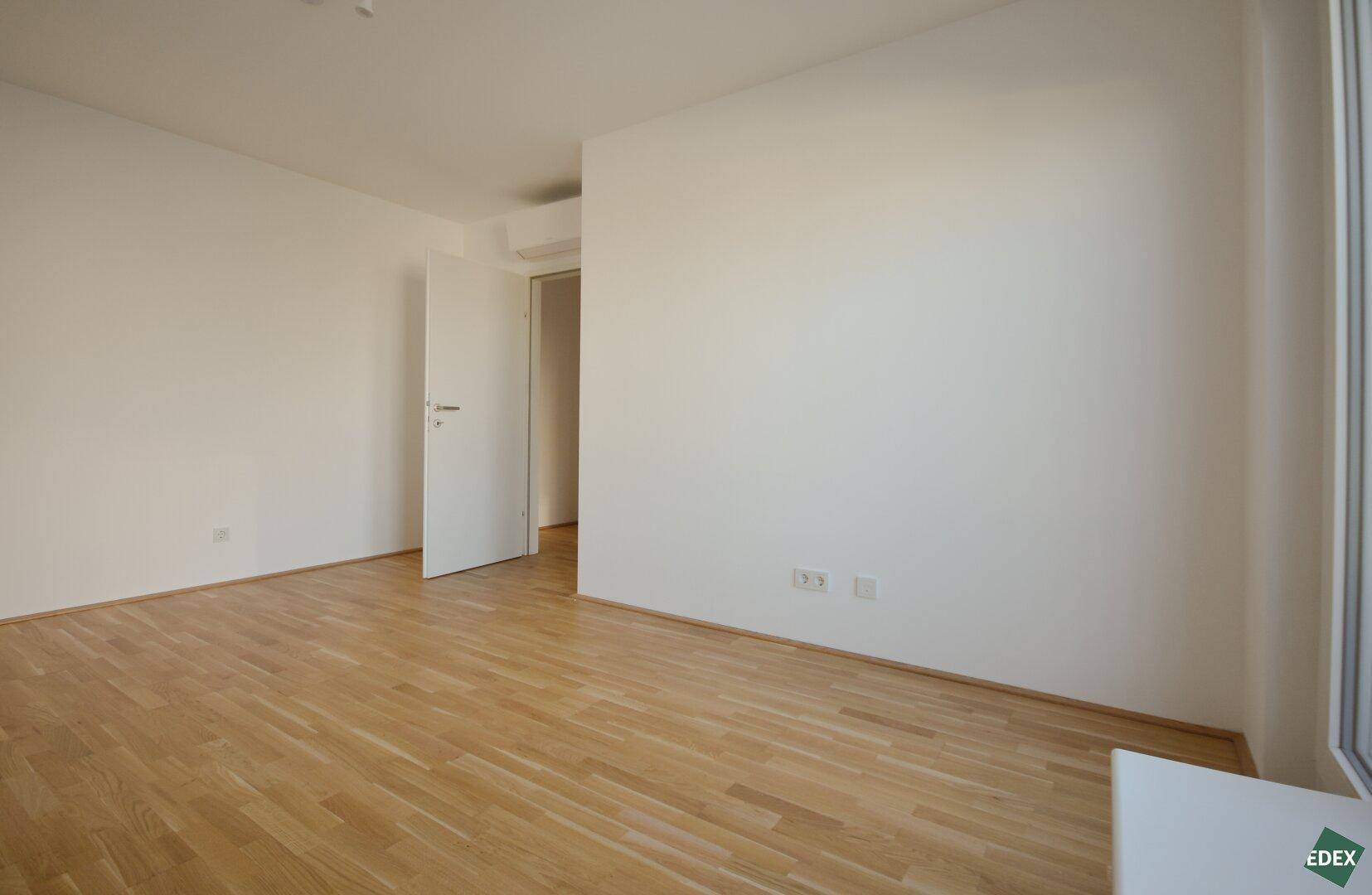 Geräumiges, leeres Schlafzimmer mit Holzboden und weiß gestrichenen Wänden, vielseitig nutzbar.