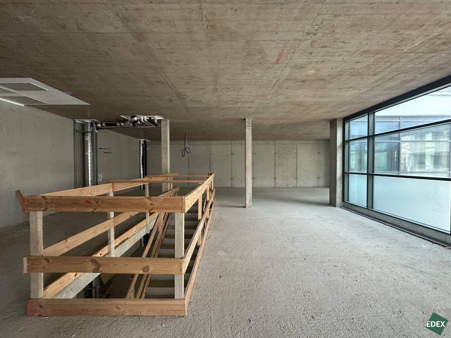 Großer Gewerberaum im Rohbau mit Betonoberflächen und einer Holzkonstruktion, die eine Rampe oder Treppe andeutet.
