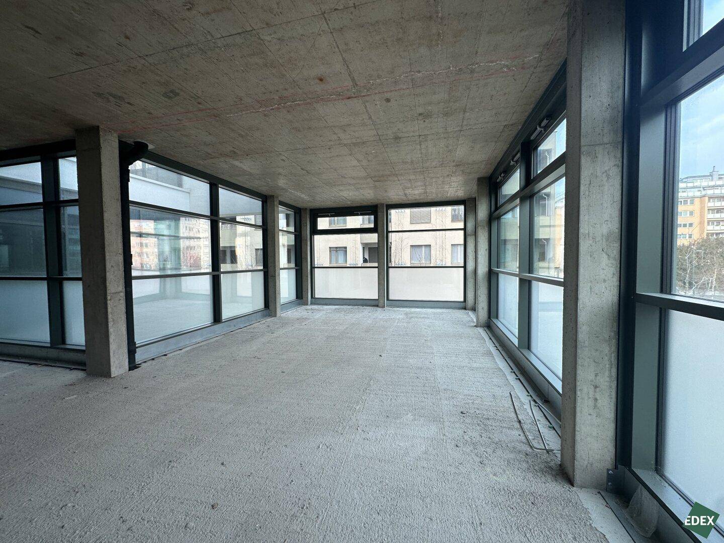 Heller Gewerberaum im Rohbau mit Betonboden und großen Fensterfronten, die viel Tageslicht hereinlassen.
