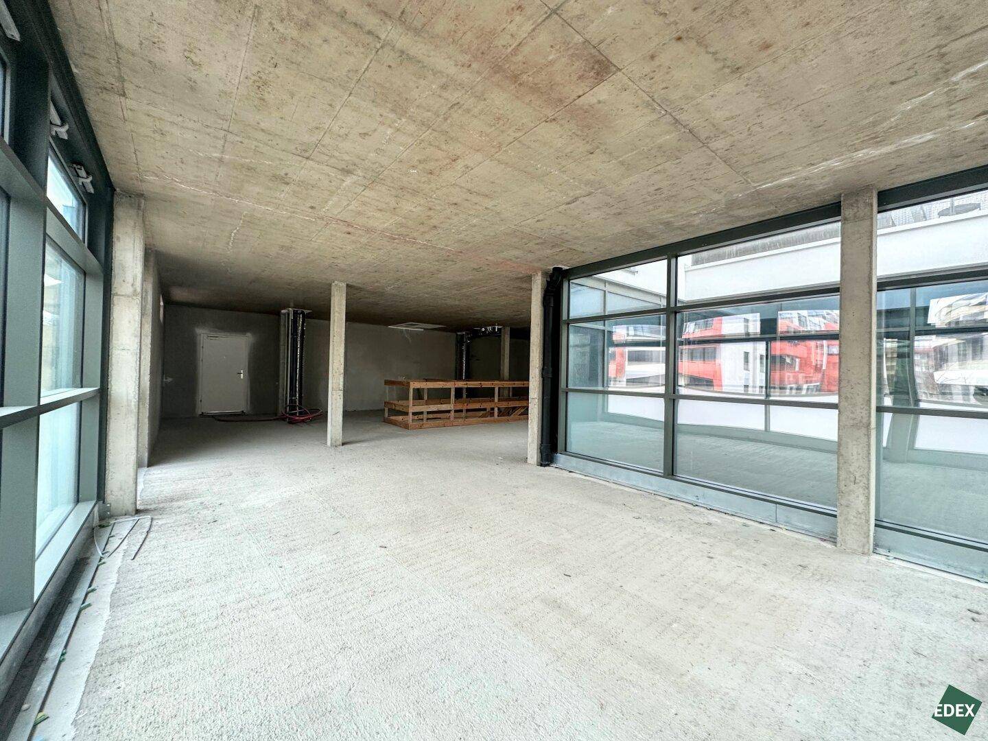 Geräumiger Gewerberaum im Rohbau mit Betonwänden und -boden, großen Fenstern und Blick auf die Umgebung.