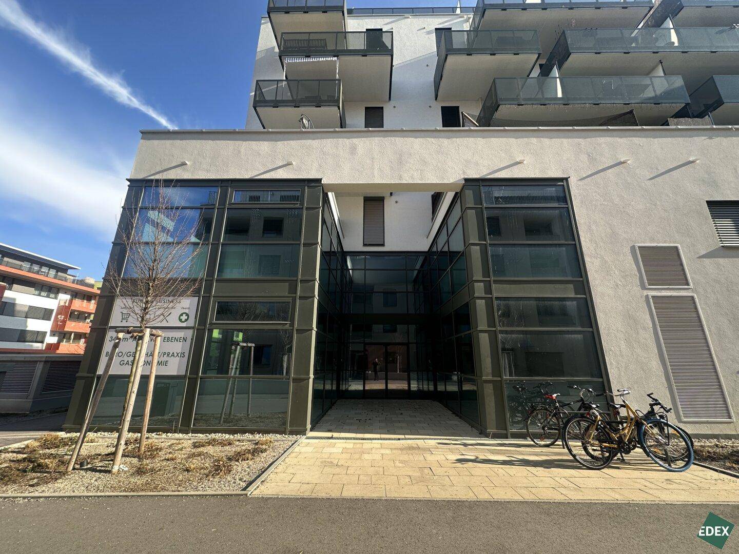 Eingangsbereich eines modernen Geschäftsgebäudes mit großen Glasfronten und Fahrradständern davor unter blauem Himmel.