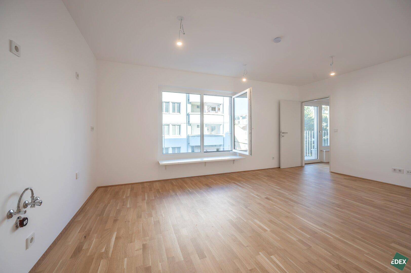 Leerer Wohnraum mit großen Fenstern, Parkettboden und Anschlussmöglichkeiten für eine Küche.