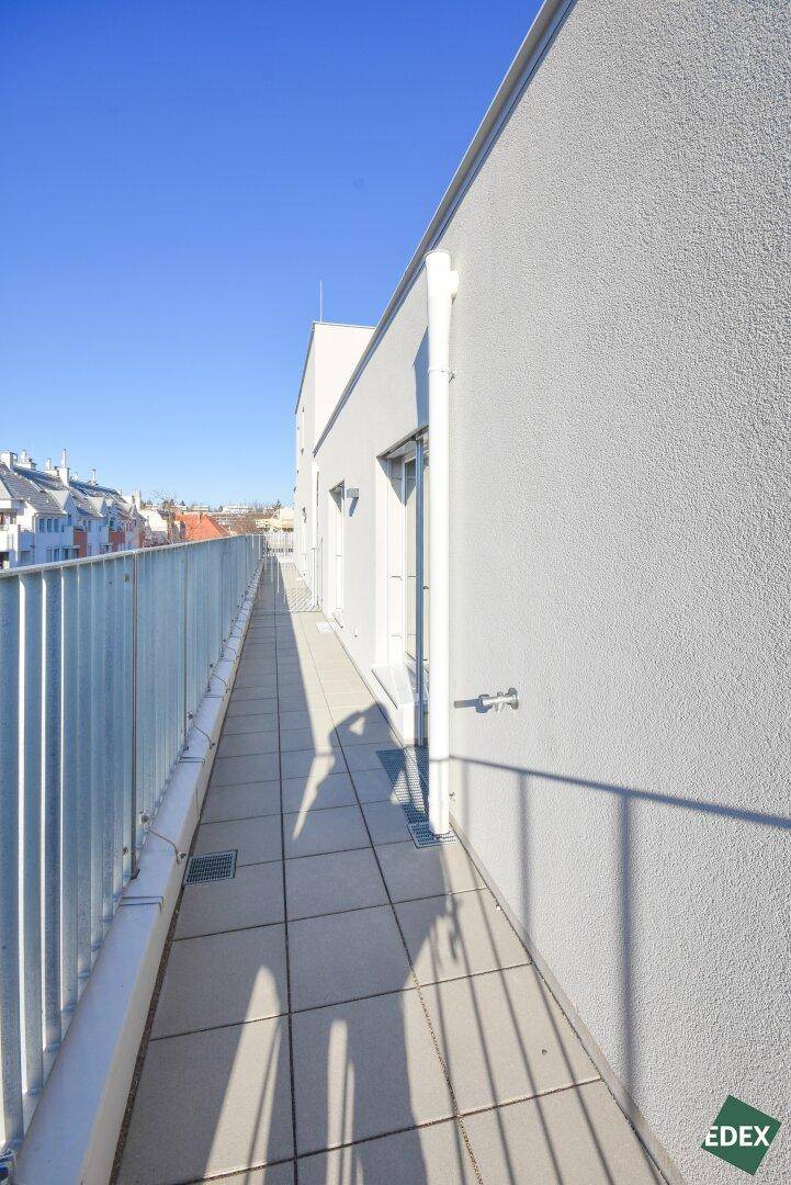 Langer, schmaler Balkon mit Fliesenboden und weißer Fassade unter blauem Himmel.