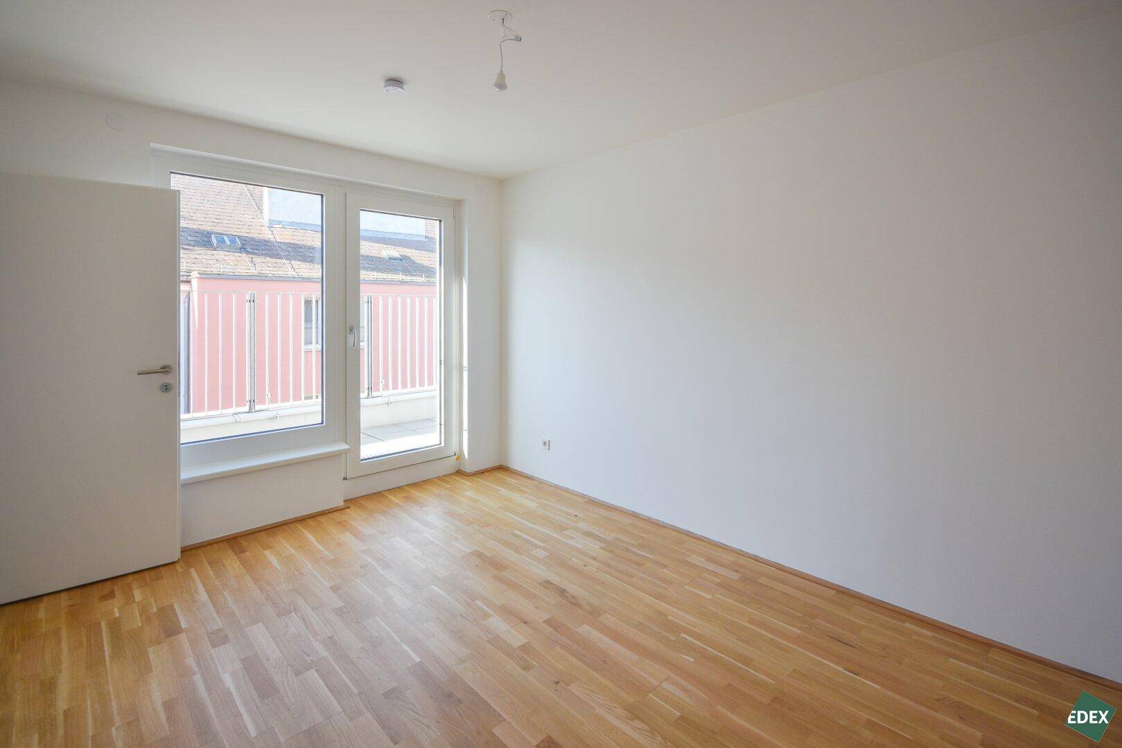 Leeres Zimmer mit hellem Parkettboden und großen Fenstern, die viel Tageslicht hereinlassen.
