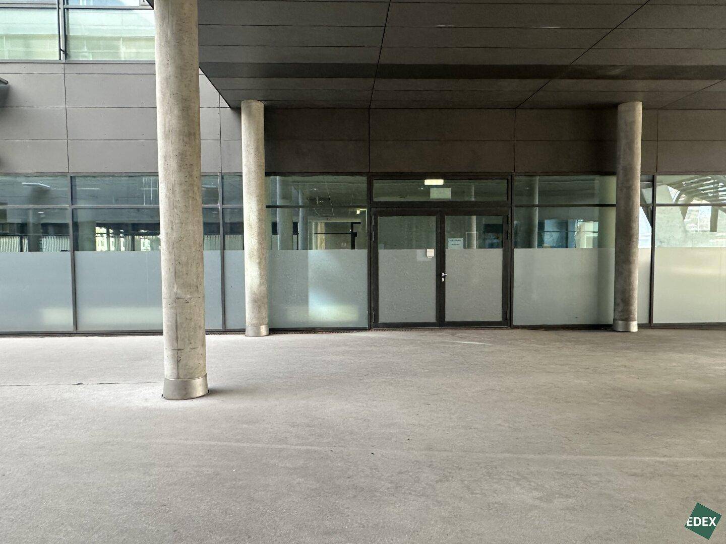 Moderner Gebäudeeingang mit großen Glasflächen, doppelten Türen und robusten Betonsäulen.