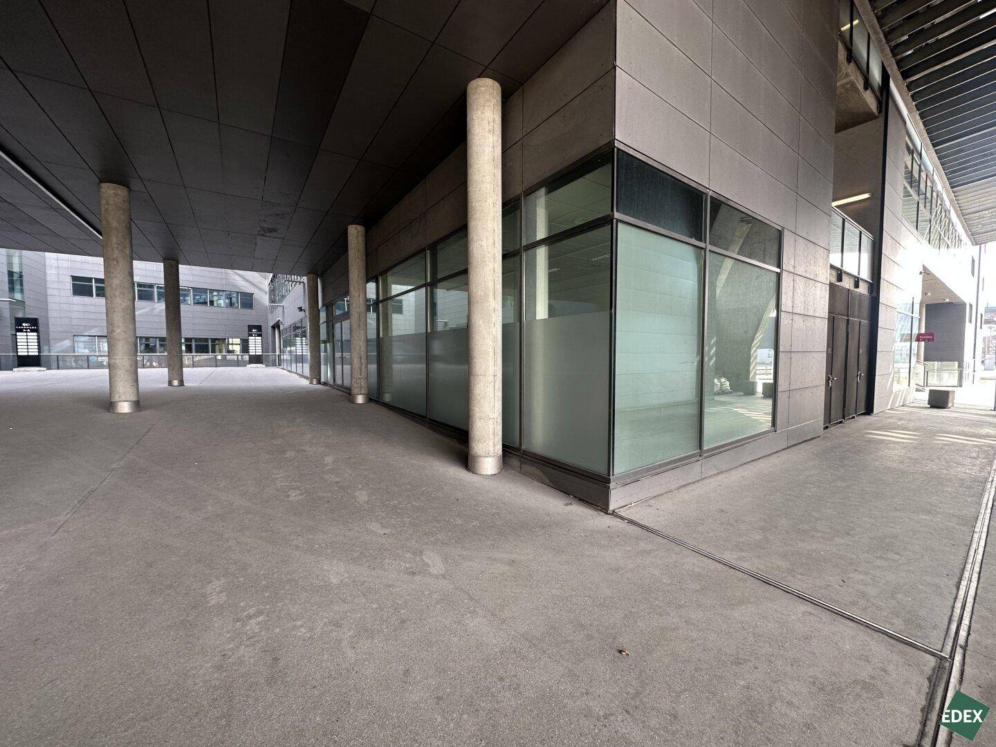 Außenansicht eines modernen Gebäudes mit Glasfassade, Betonsäulen und einem breiten Gehweg.