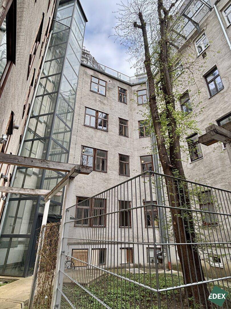 Innenhof eines Gebäudes mit modernem Glasaufzug und alten Bäumen.