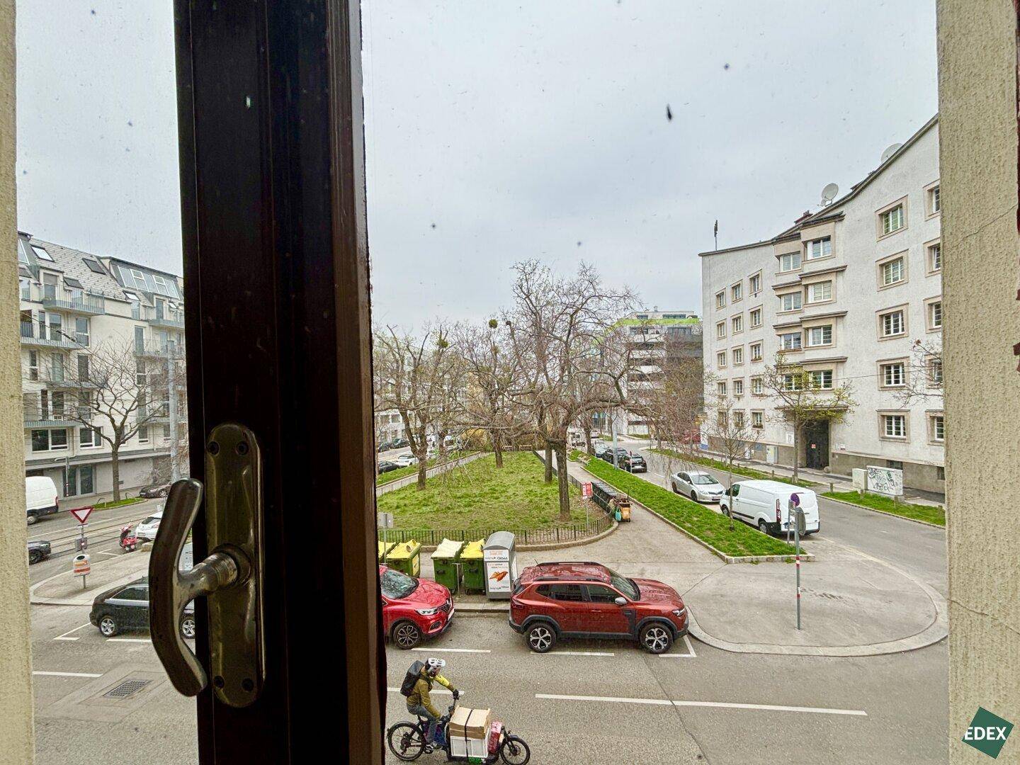 Blick aus dem Fenster auf eine belebte Straße mit Grünfläche und umliegenden Gebäuden.