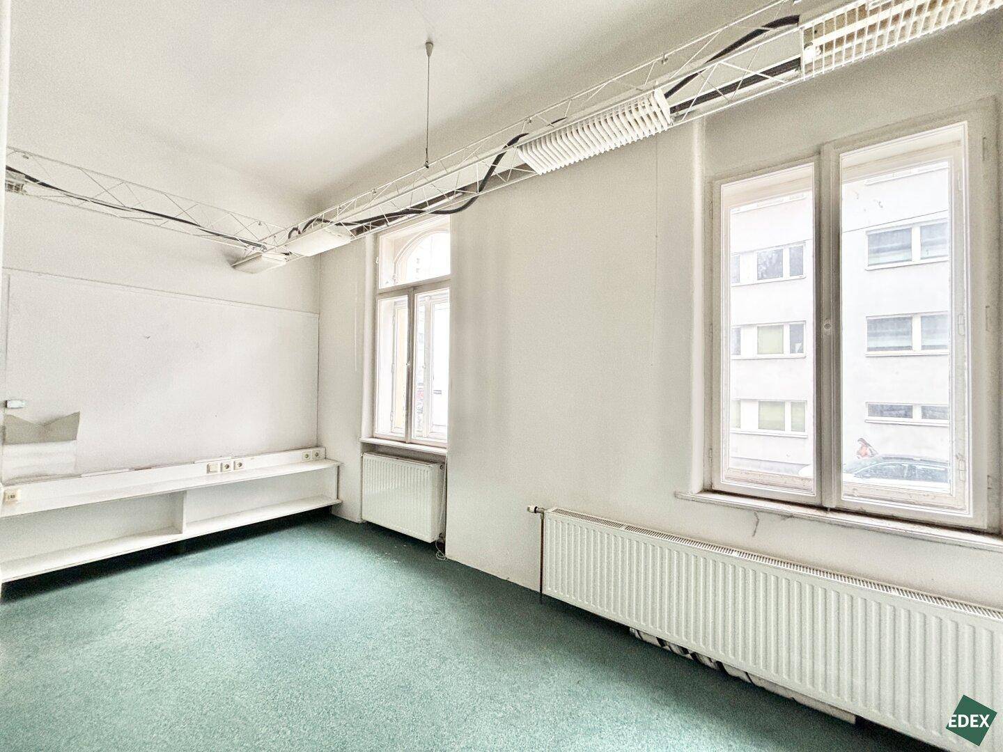 Heller Büroraum mit großen Fenstern, Heizkörpern und einfacher Beleuchtung.