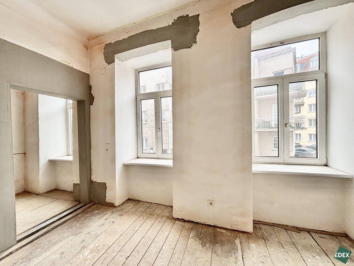 Leerer Raum mit abgenutzten Holzdielen, unfertigen Wänden und zwei großen Fenstern, die viel Licht hereinlassen.