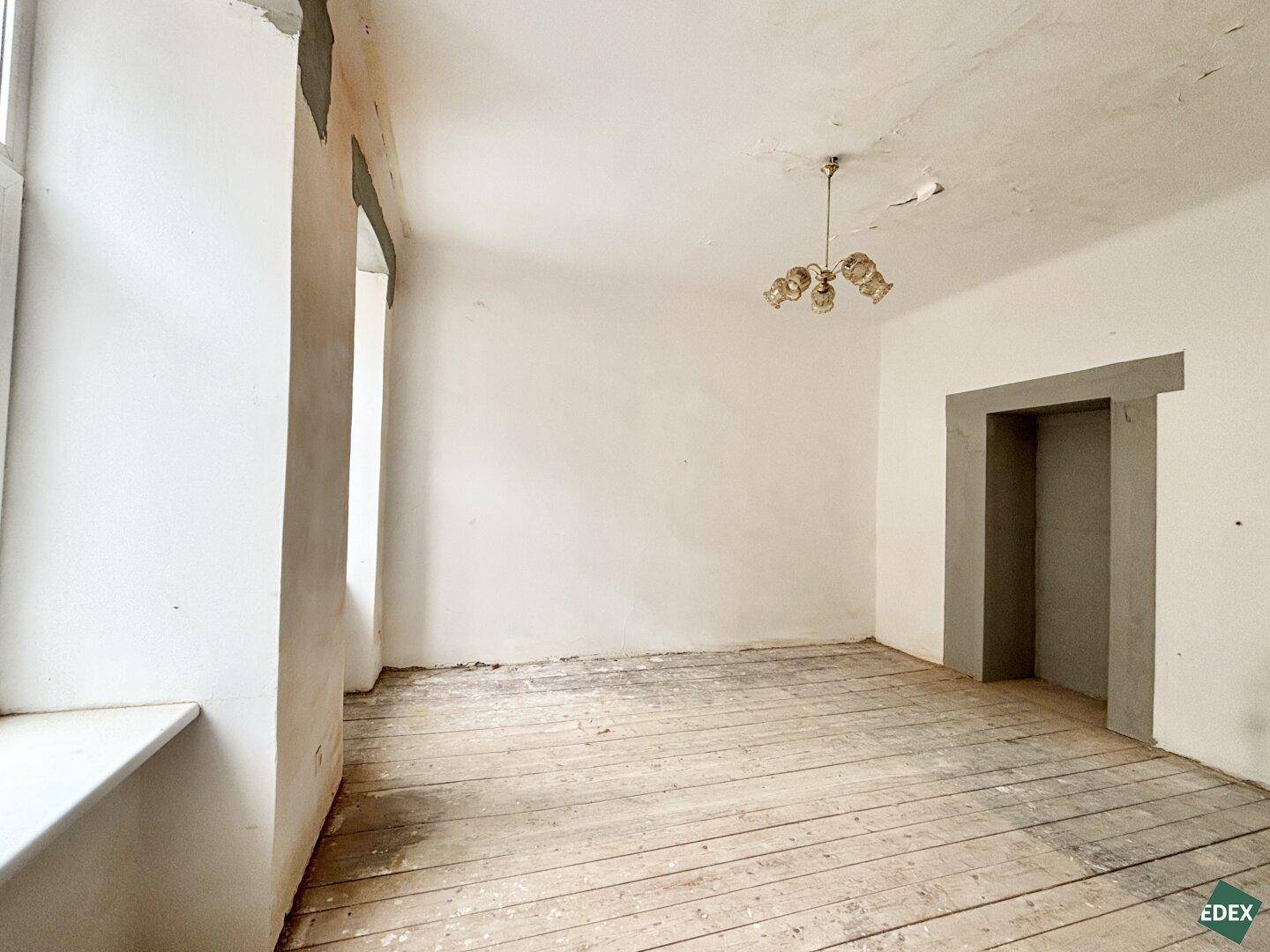 Unmöblierter Raum mit abgenutzten Holzdielen, hohen Decken und einem Fenster, das auf Renovierung wartet.