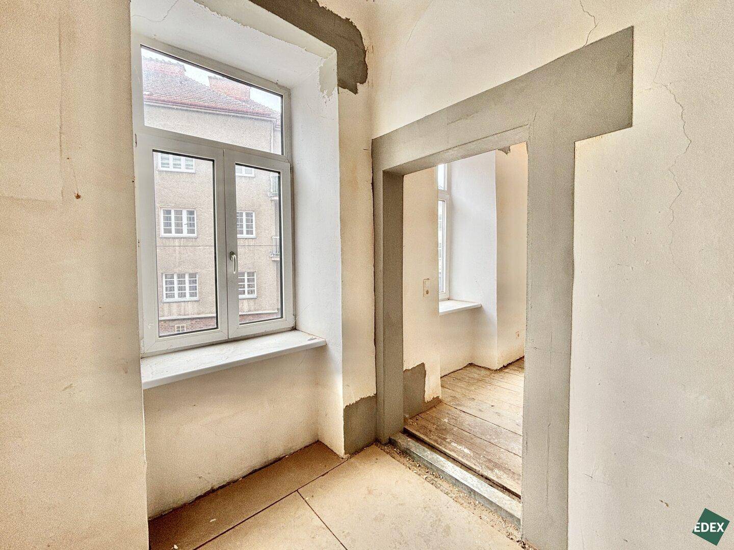 Kleiner, unfertiger Raum mit einem Fenster und einem Durchgang zu einem weiteren Bereich, der renoviert werden muss.