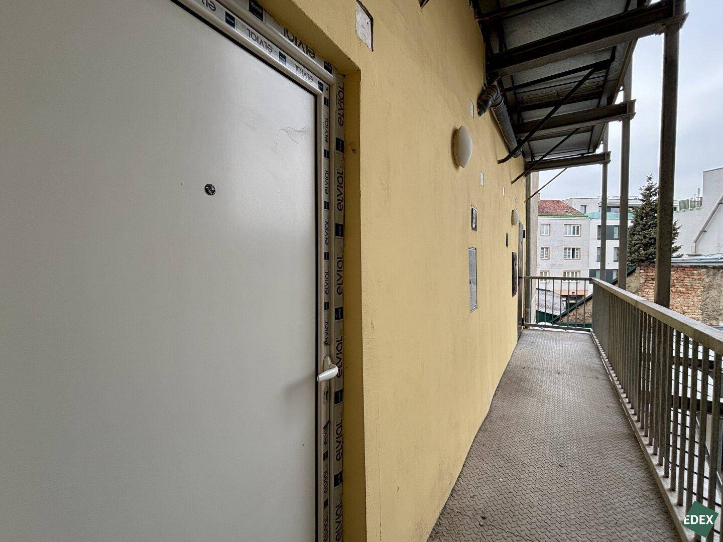 Langer Außenbalkon mit Metallgeländer und einer weißen Tür, der Zugang zu den Wohnungen bietet.