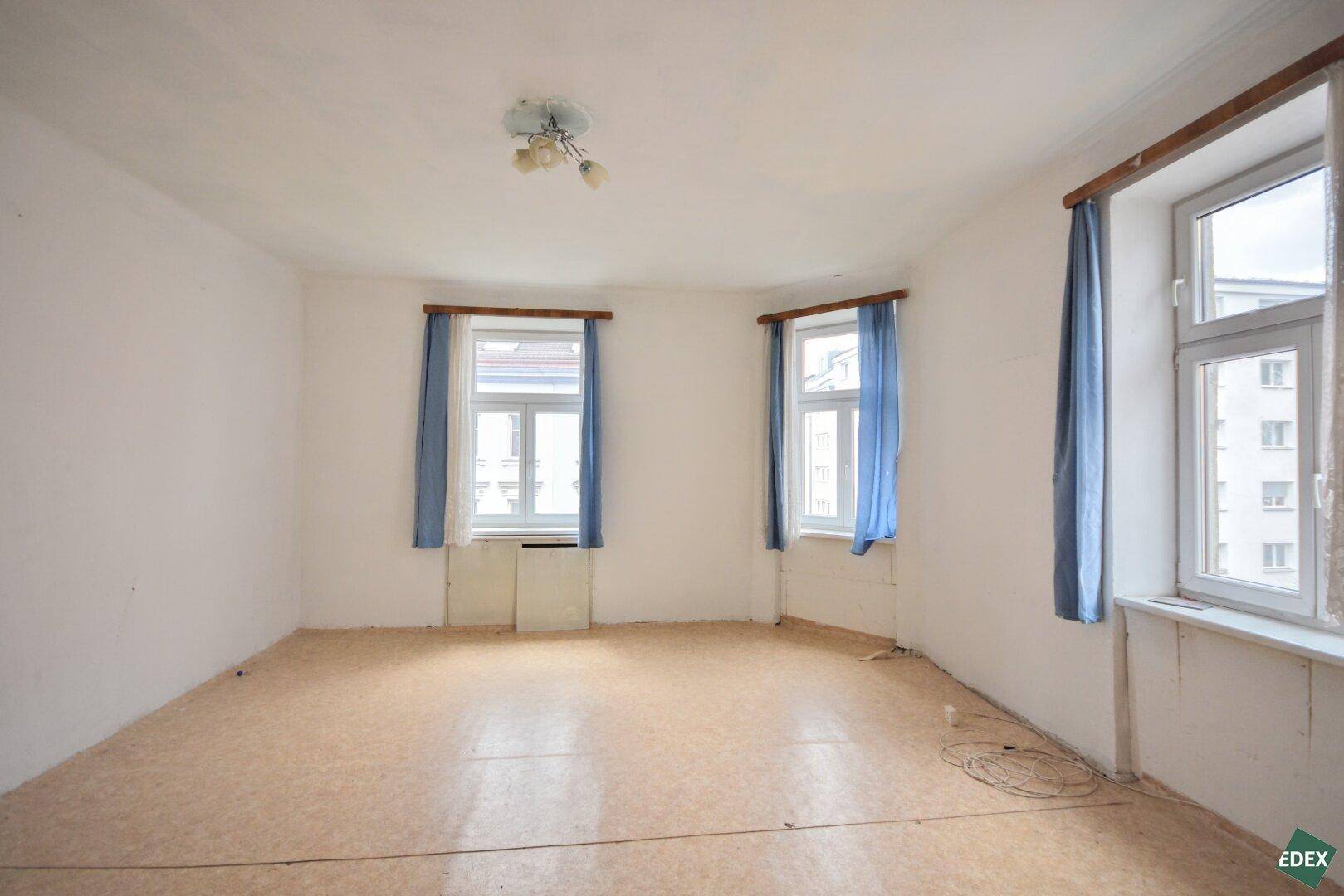 Geräumiges Zimmer mit Linoleumboden und drei Fenstern, das auf eine Modernisierung wartet.