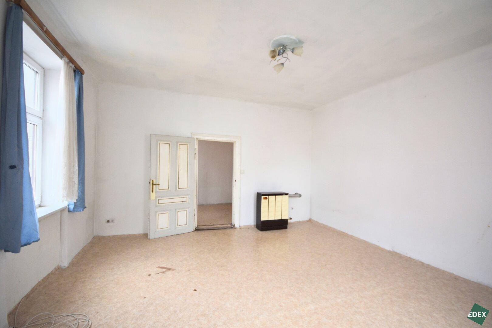 Leeres Zimmer mit Linoleumboden, zwei Fenstern und einer alten Tür, die in einen weiteren Raum führt.