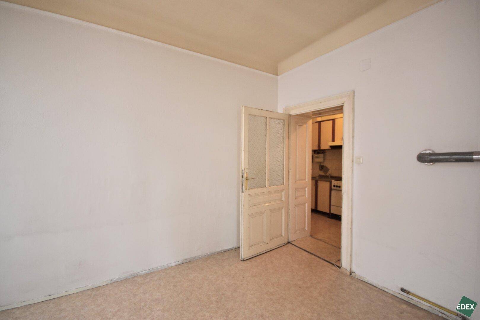 Leeres Zimmer mit Linoleumboden und einer Tür, die den Blick in eine kleine Küche freigibt.