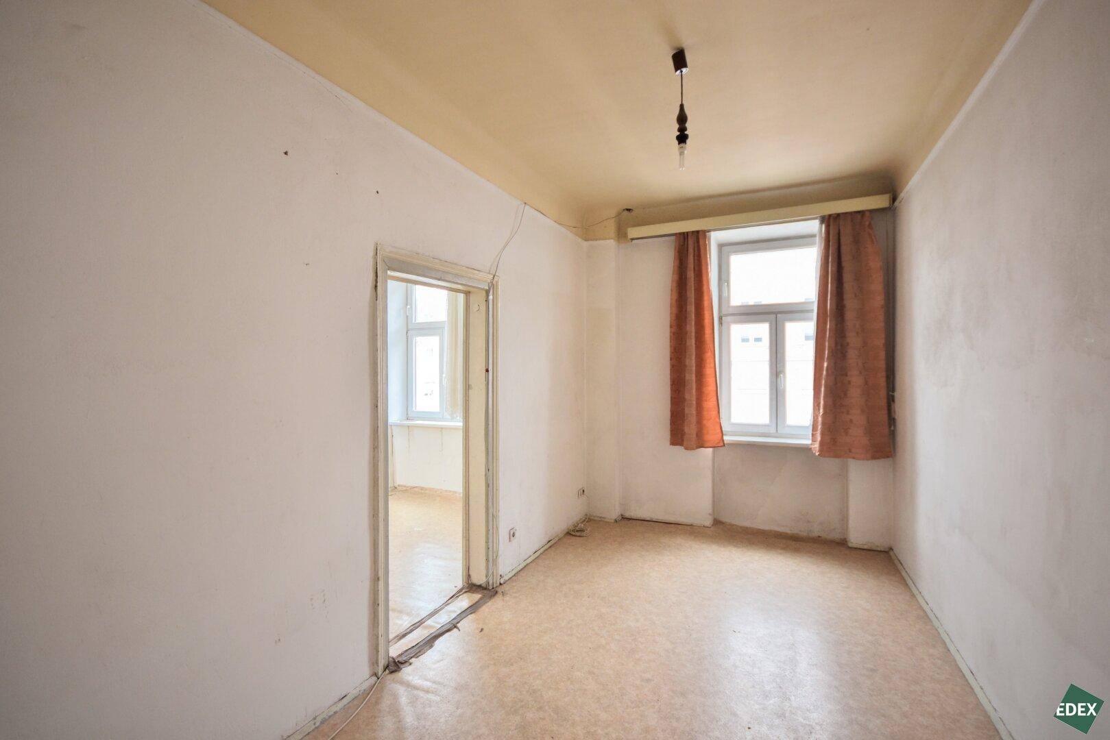 Einfaches Zimmer mit Linoleumboden, einem Fenster und einer Tür, die in einen anderen Raum führt.