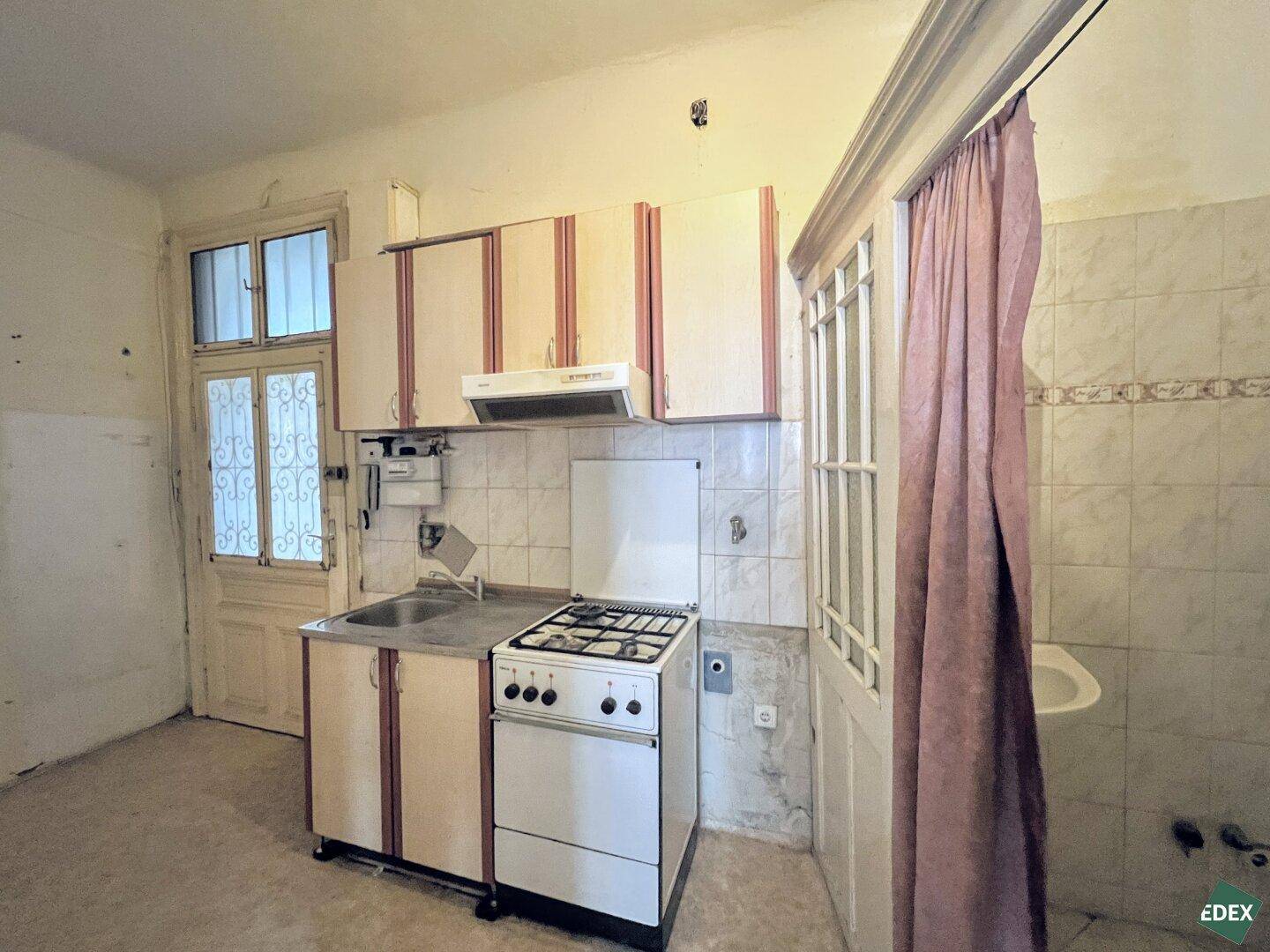 Funktionale, aber renovierungsbedürftige Küche mit Gasherd und Spüle, mit Blick auf eine Duschkabine.