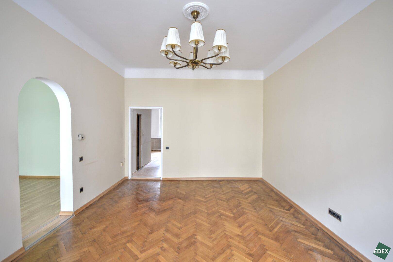 Helles, leeres Zimmer mit Fischgrätparkett und einem eleganten Rundbogen zu einem weiteren Raum.
