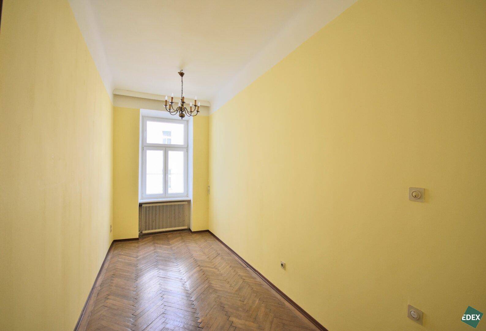 Schmales, sonniges Zimmer mit gelben Wänden, Fischgrätparkett und einem Fenster.