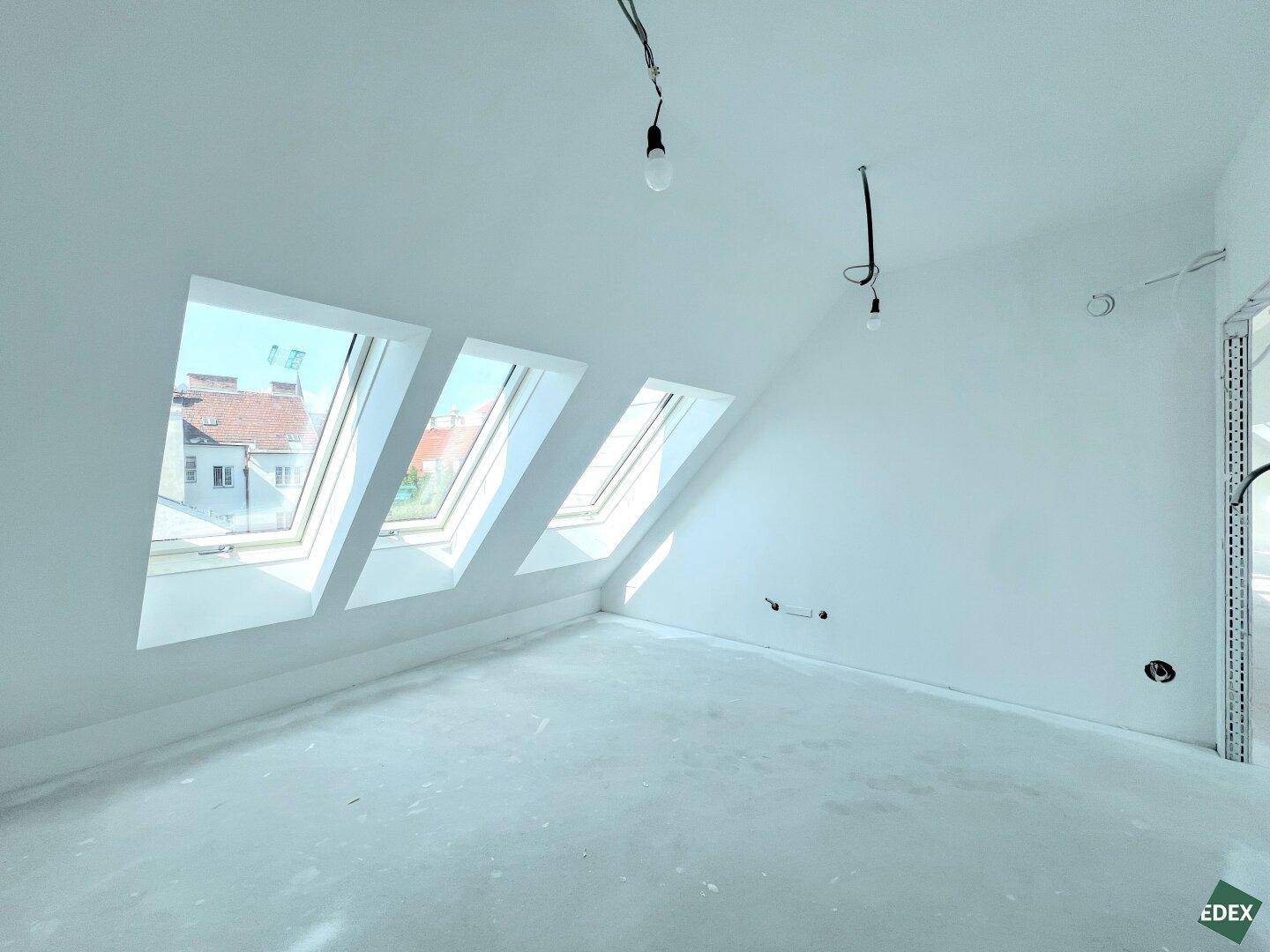 Heller Rohbau im Dachgeschoss mit mehreren Dachfenstern und Blick auf die umliegenden Dächer.
