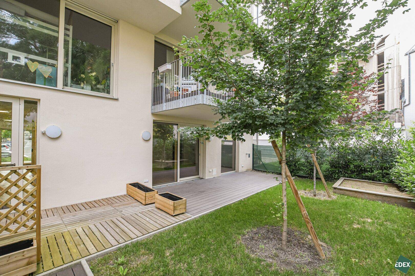 Außenbereich mit Holzterrasse, Rasen und Sandkasten, umgeben von Bäumen und Sträuchern.