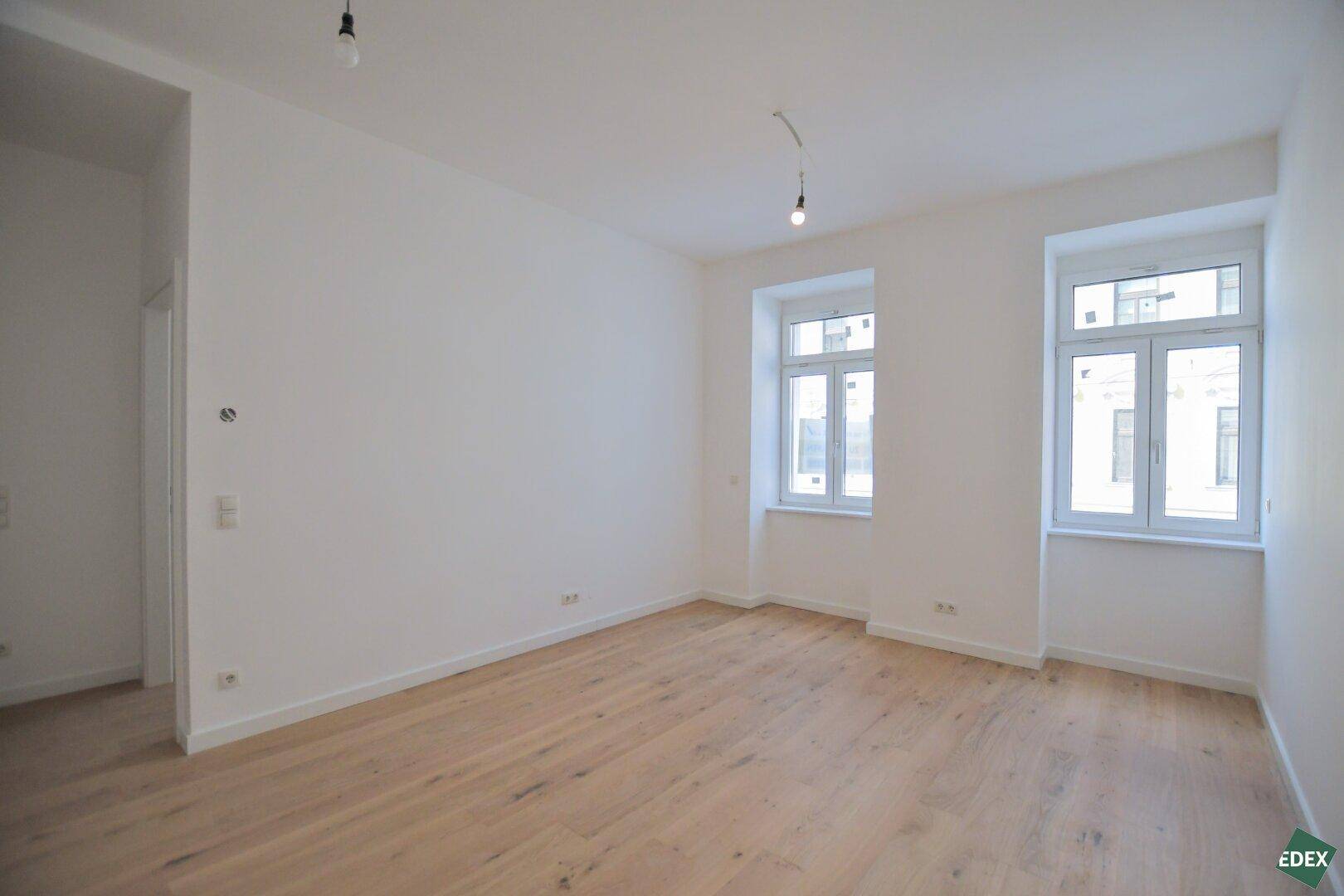 Heller, leerer Raum mit zwei großen Fenstern und Holzboden, ideal für Wohn- oder Schlafbereich.