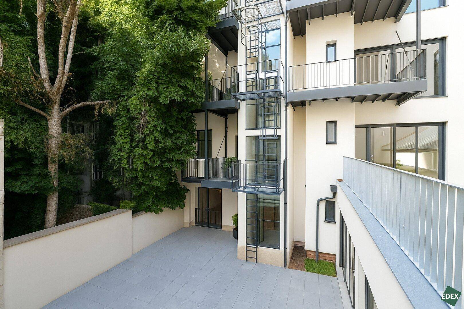 Innenhof mit externem Glasaufzug, gepflasterter Fläche und üppiger Begrünung.