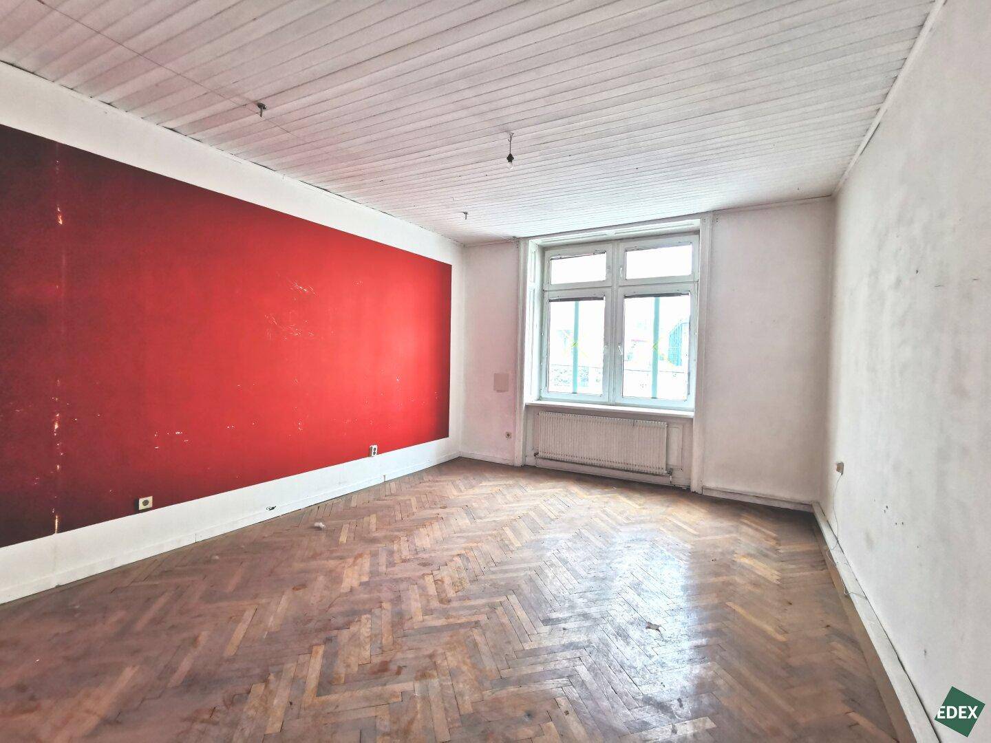 Leerer Raum mit roter Akzentwand und Fischgrätparkett, das eine Renovierung benötigt.