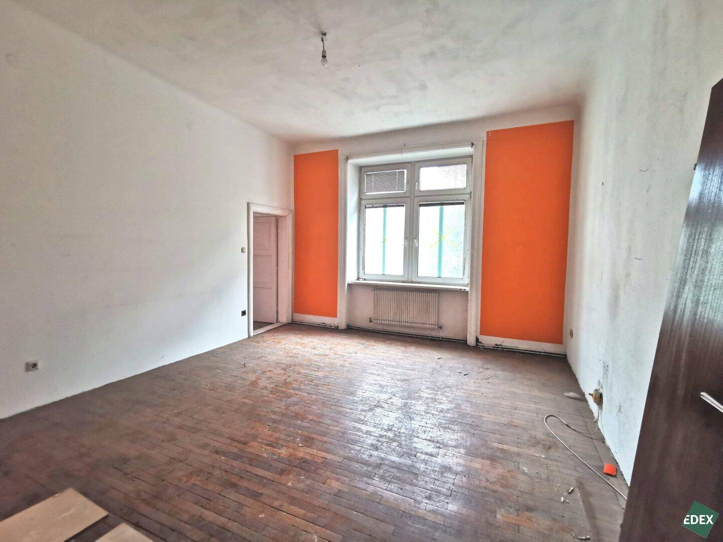 Leerer Raum mit orangefarbenen Akzentwänden und abgenutztem Holzfußboden, bereit zur Renovierung.