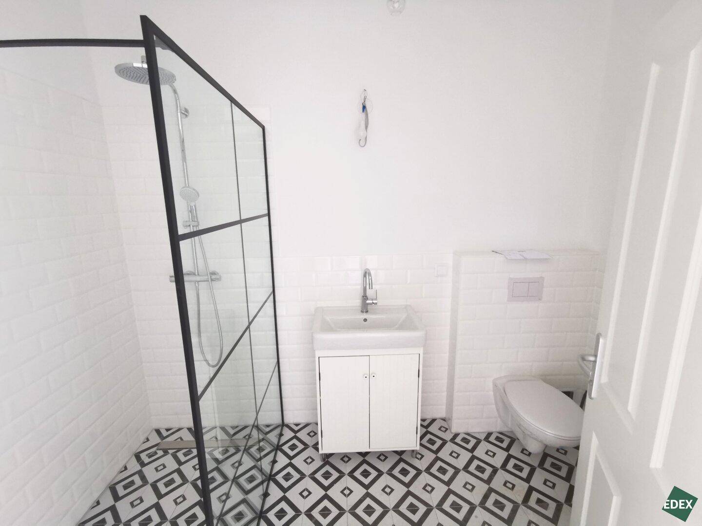 Modernes Badezimmer mit begehbarer Dusche, weißen Fliesen und auffälligem Bodenmuster.