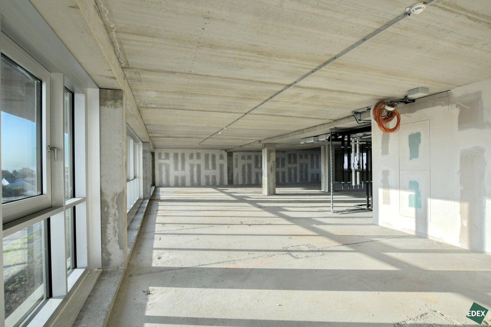 Unfertiger Büroraum mit Betonboden und großen Fenstern, die viel natürliches Licht in den Raum bringen.