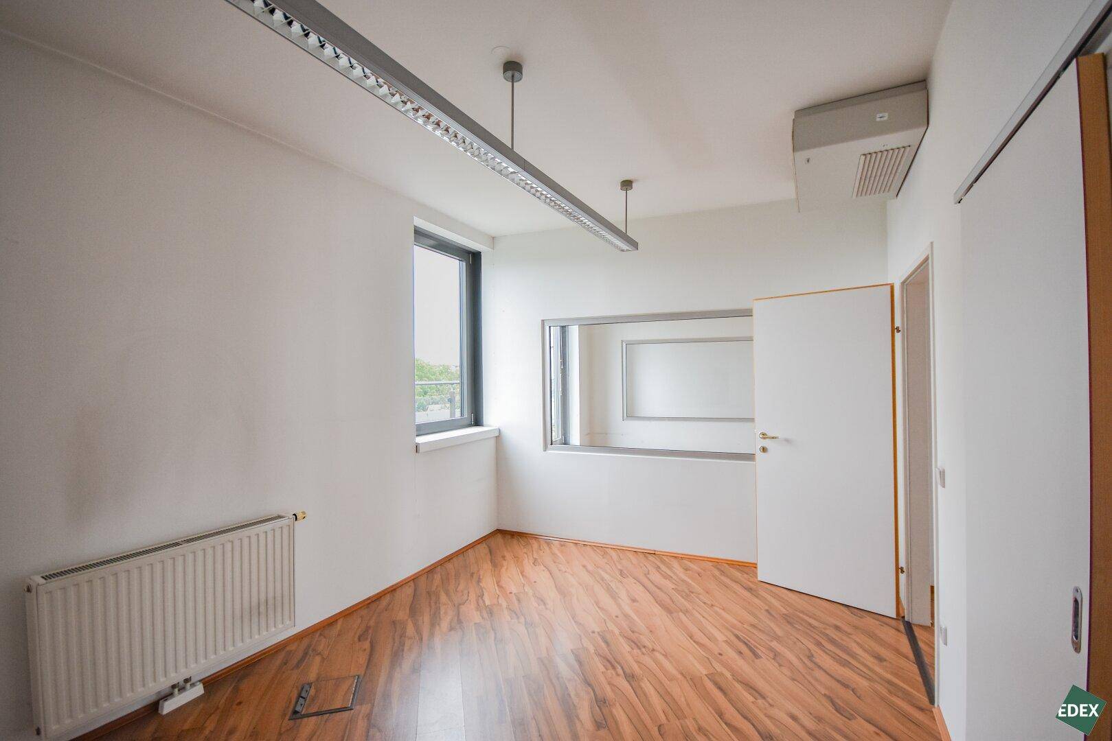 Leeres Büro mit Laminatboden, einem Fenster und einer internen Glaswand.
