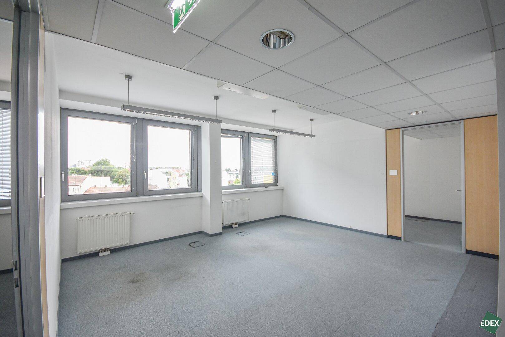 Helles Büro mit Teppichboden, großen Fenstern und Zugang zu einem weiteren Raum.