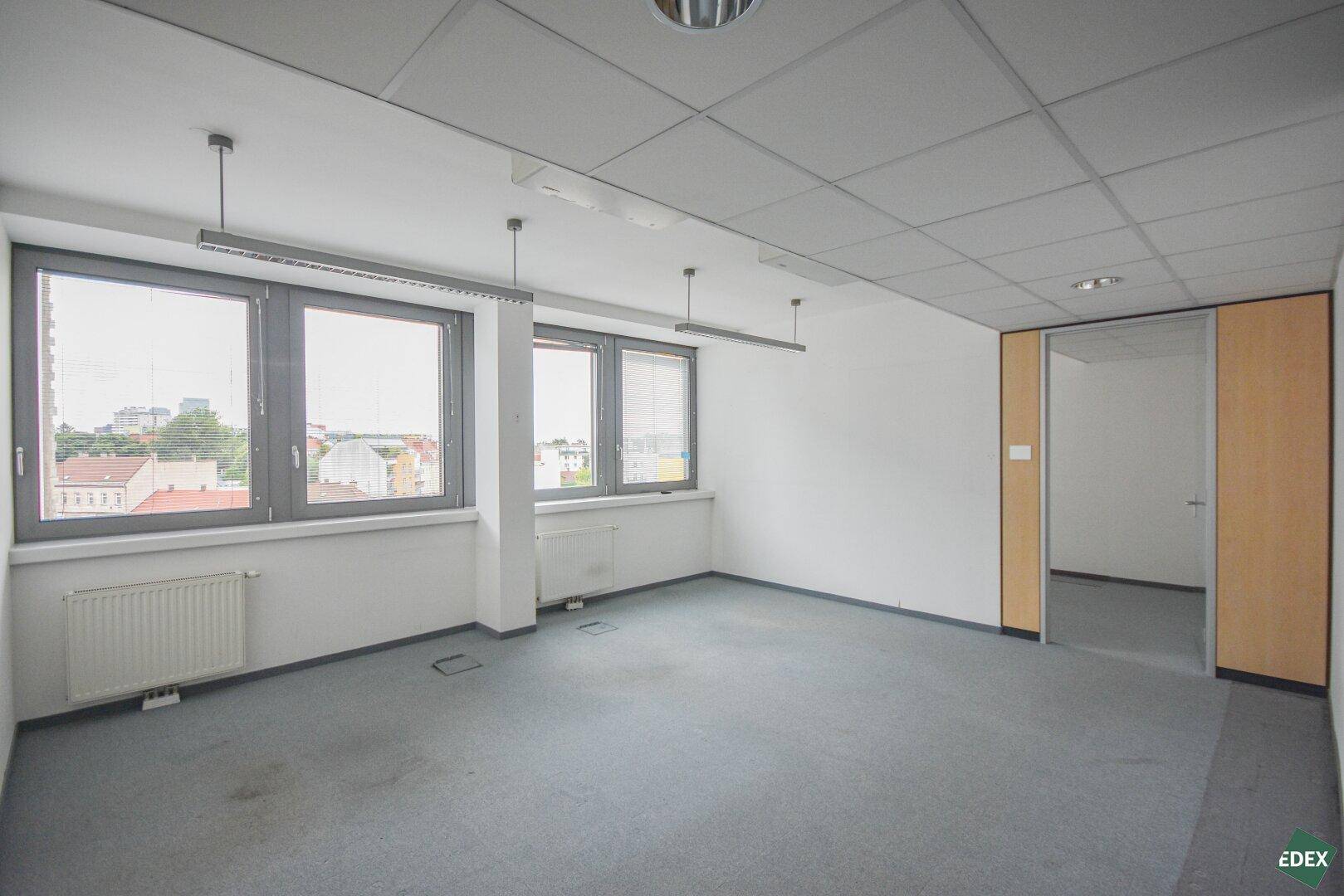 Leeres Büro mit Teppichboden, großen Fenstern und Blick auf die Stadt.