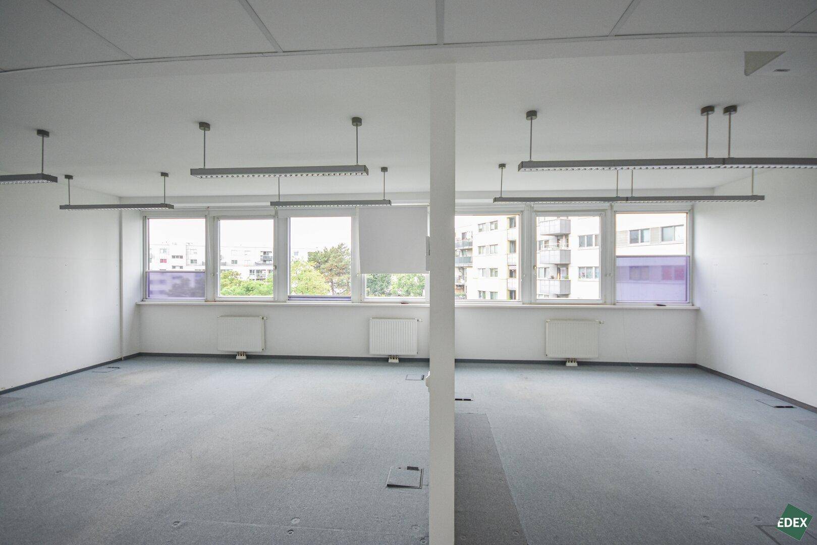 Helles Büro mit Teppichboden, vielen Fenstern und Blick auf umliegende Gebäude.
