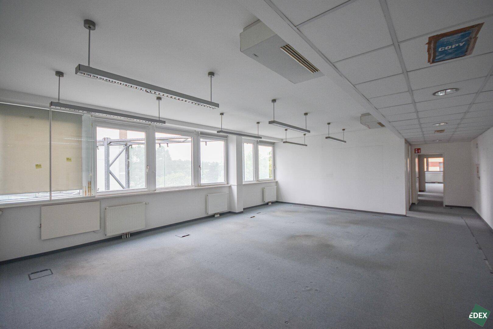 Leeres Büro mit Teppichboden, großen Fenstern und Blick auf die umliegenden Gebäude.