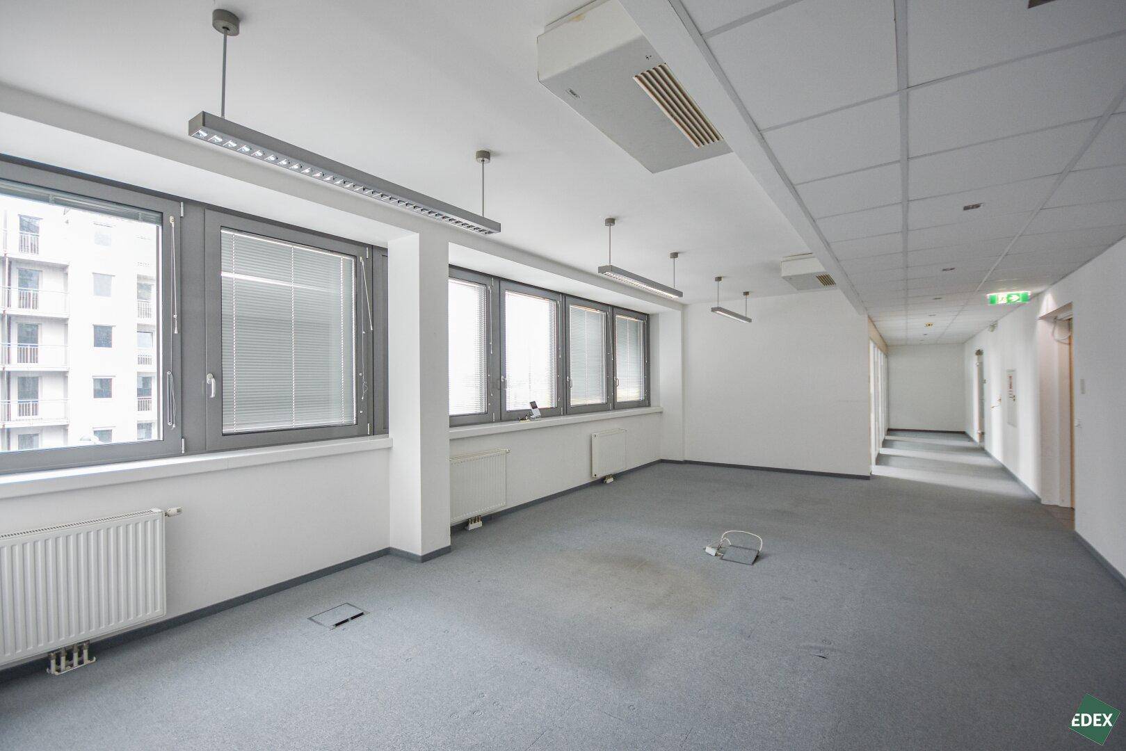 Geräumiges Büro mit Teppichboden, großen Fenstern und Klimaanlage an der Decke.