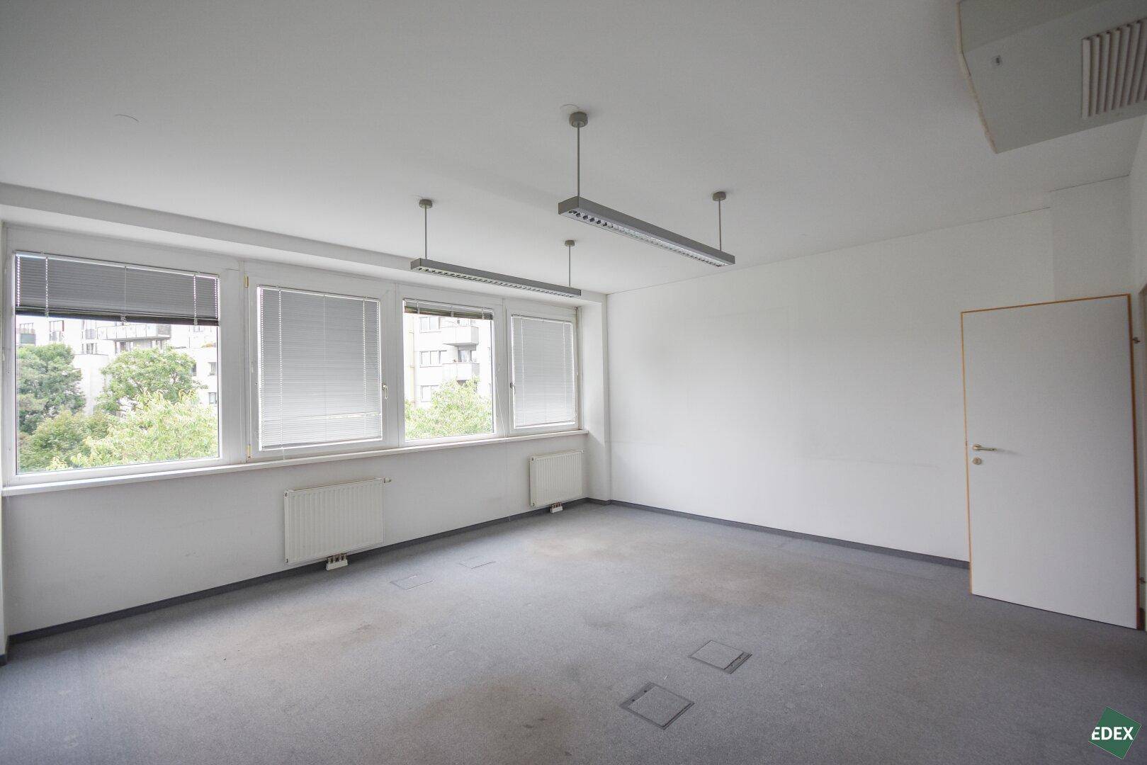 Leeres Büro mit Teppichboden, weißen Wänden, Jalousien an den Fenstern und einer Tür.