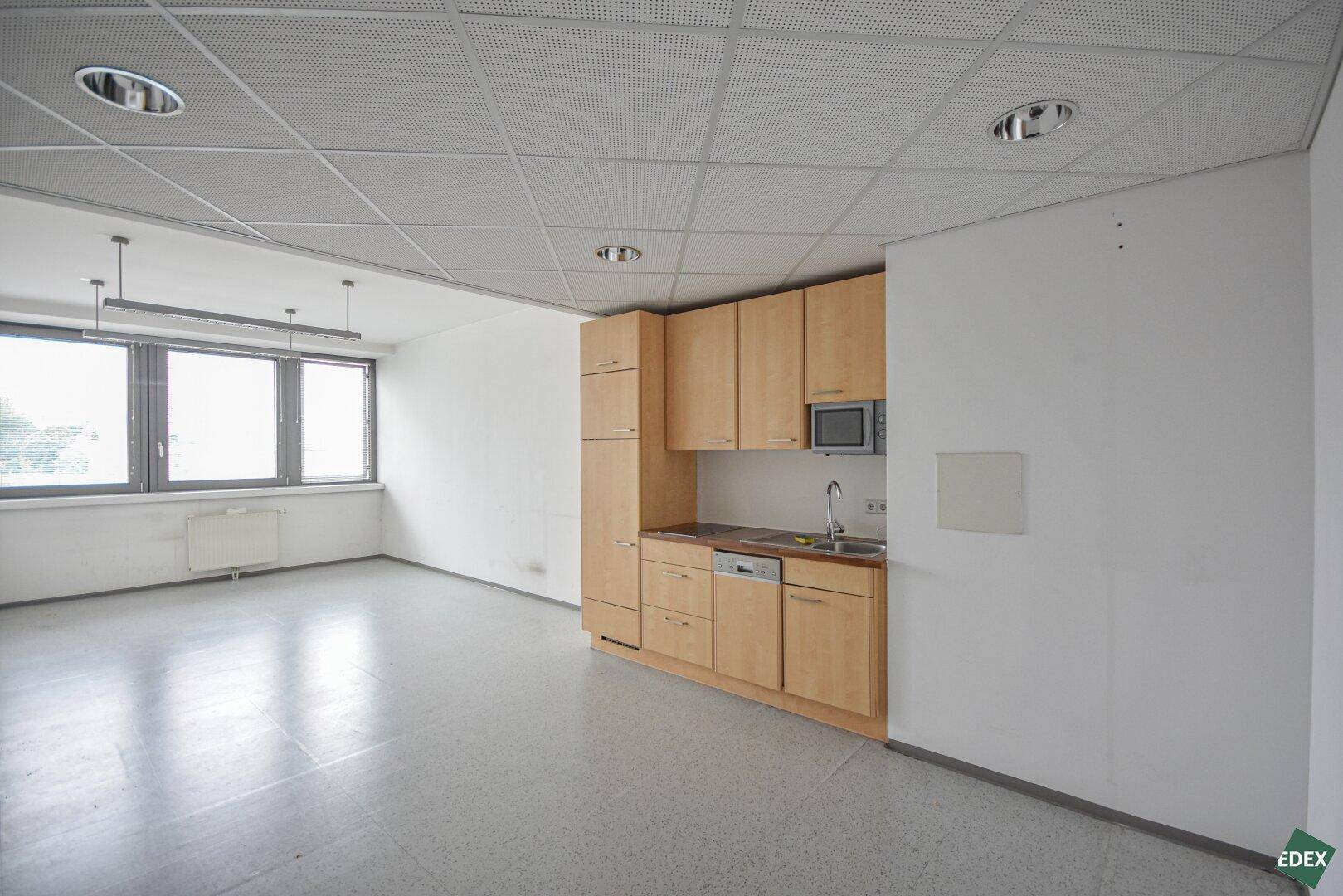 Heller Büroraum mit integrierter Küchenzeile, Rasterdecke und großen Fenstern.
