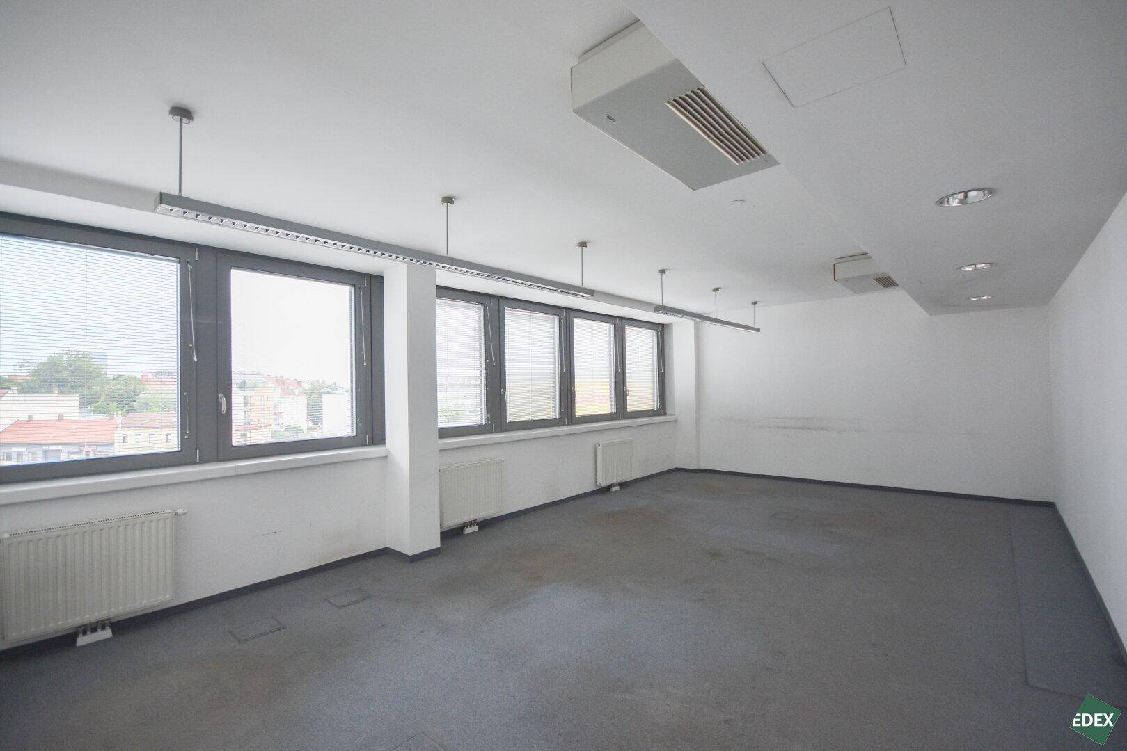 Helles und geräumiges Büro mit Teppichboden, großen Fenstern und Deckenbeleuchtung.