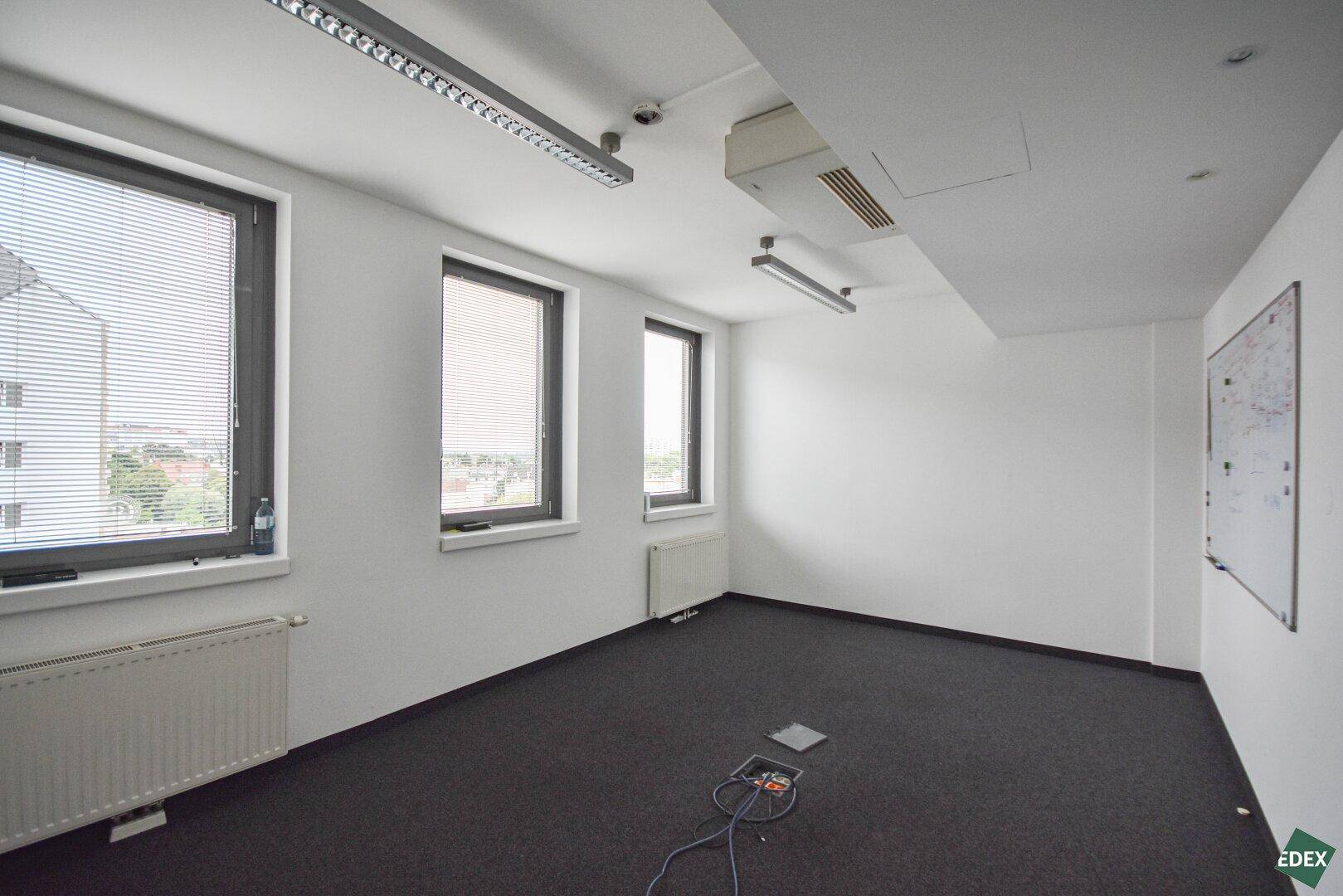 Leerer Büroraum mit Teppichboden, Fenstern mit Jalousien und Heizkörpern, bietet viel Tageslicht.
