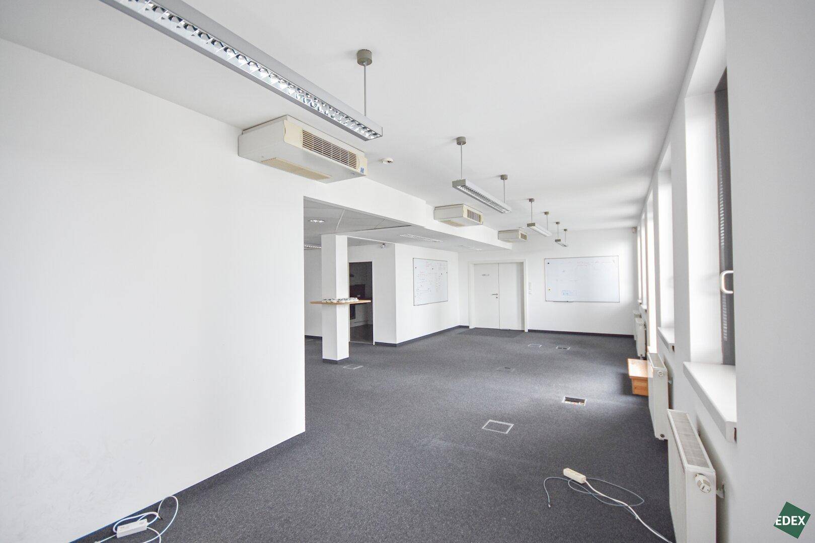 Helles Büro mit Teppichboden, Whiteboards und einem kleinen Barbereich im Hintergrund, ideal für Besprechungen.
