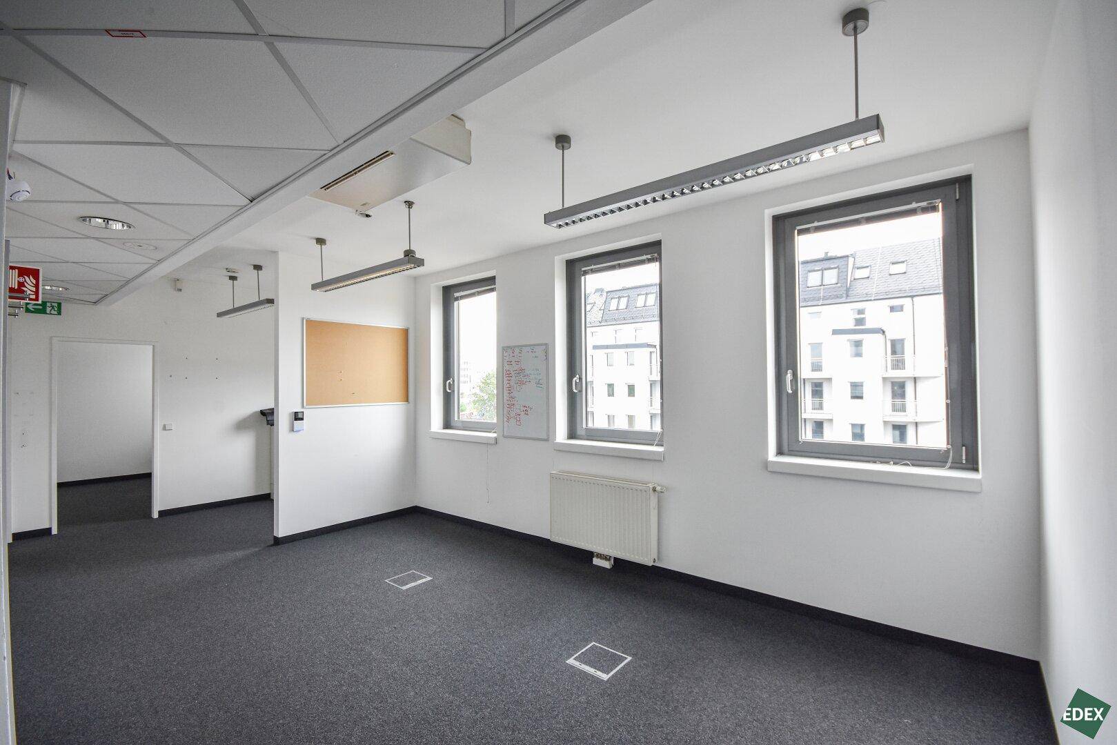 Helles Büro mit Teppichboden, großen Fenstern und einer Korktafel, ideal für kreative Arbeitsbereiche.