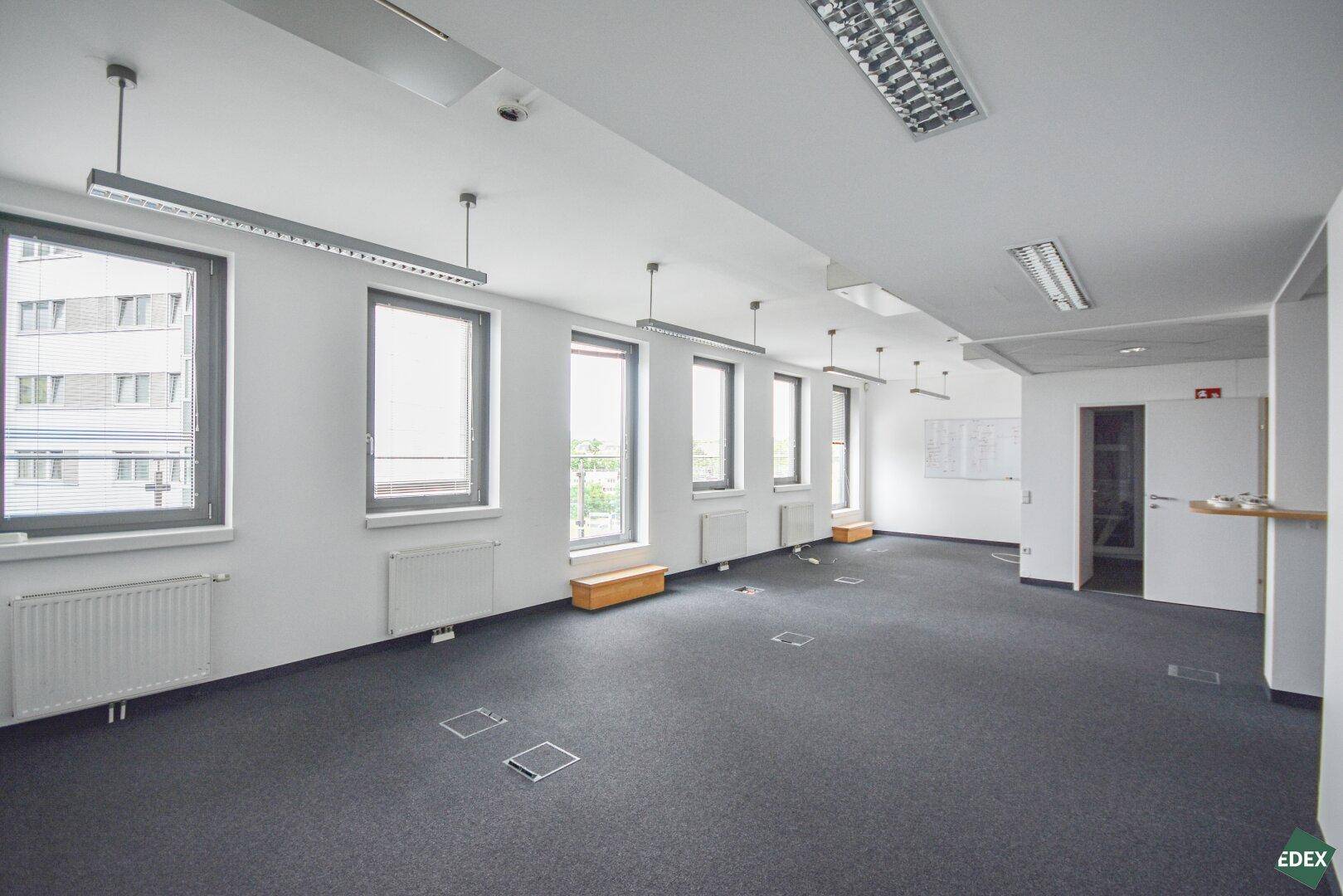 Großzügiger, leerer Büroraum mit vielen Fenstern, Teppichboden und Bodentanks für flexible Arbeitsplatzgestaltung.