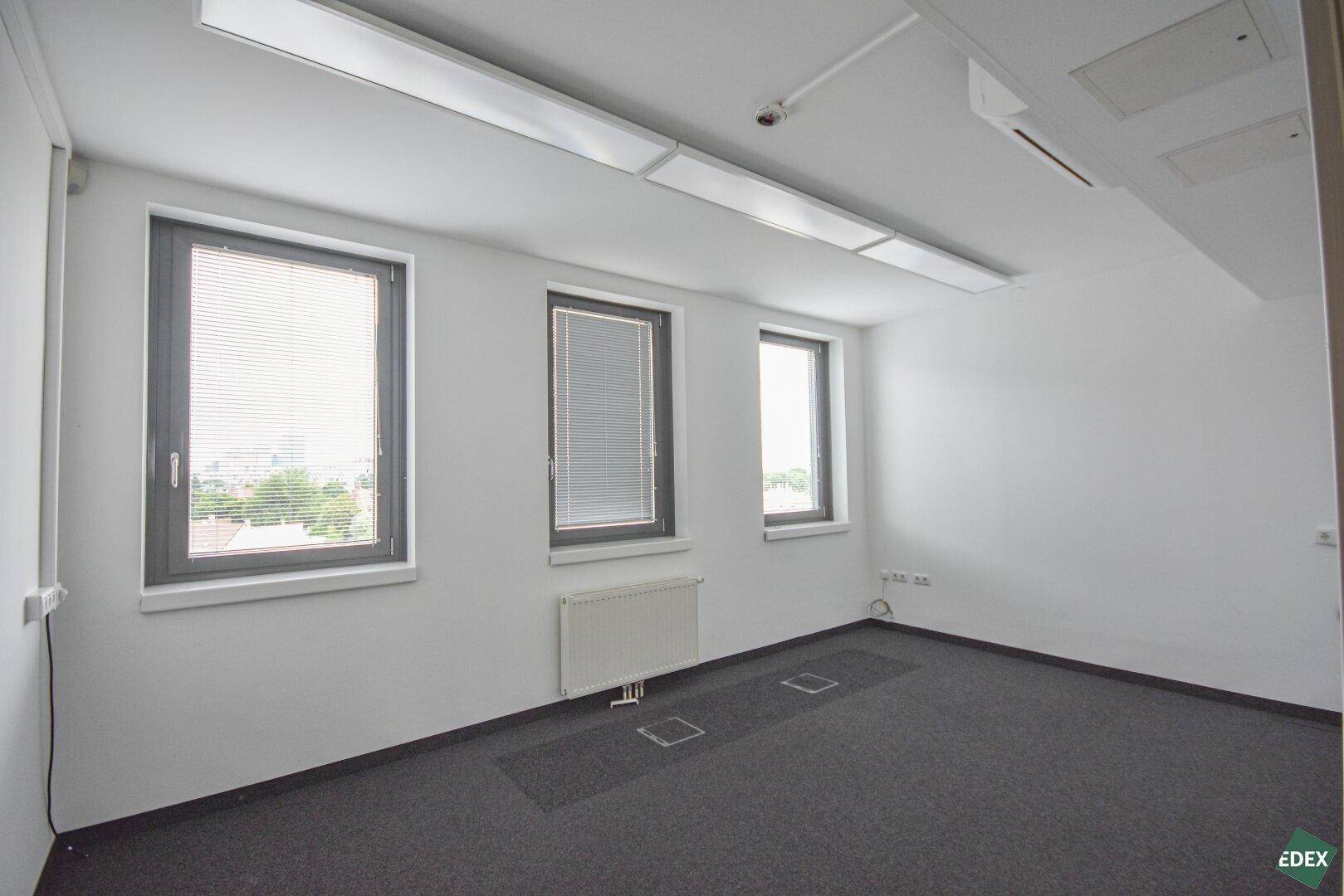 Kleinerer Büroraum mit Teppichboden, drei Fenstern mit Jalousien und Heizkörper, bietet ruhige Arbeitsatmosphäre.