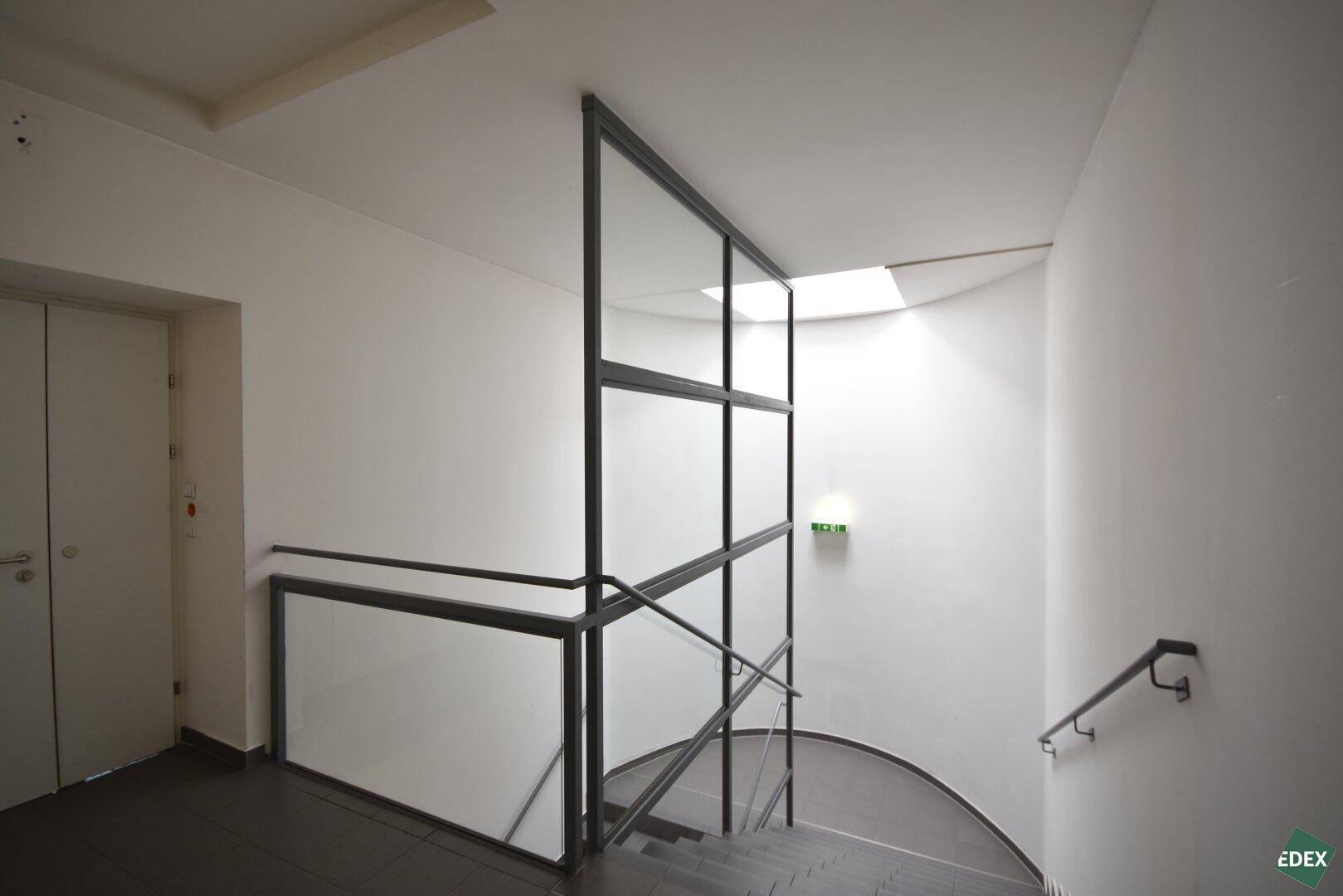 Modernes Treppenhaus mit Glas- und Metallgeländer sowie einem Oberlicht, das für Helligkeit sorgt.
