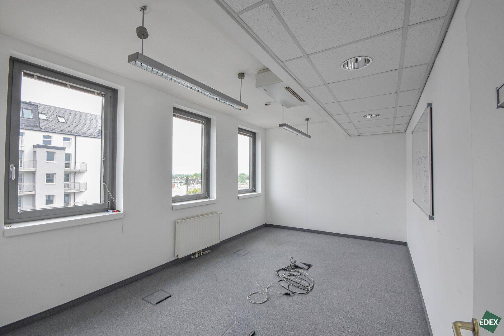Helles Büro mit Teppichboden, drei Fenstern und Blick auf umliegende Gebäude, bietet gute Aussicht.