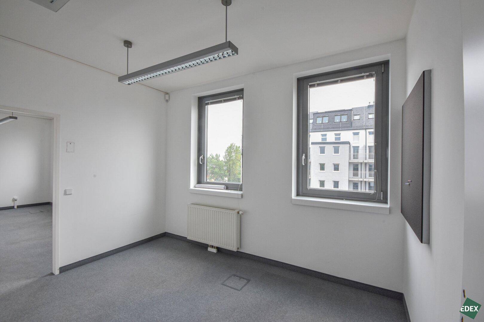 Kleineres Büro mit Teppichboden, zwei Fenstern und Blick auf die Stadt, ideal für Einzelbüros.